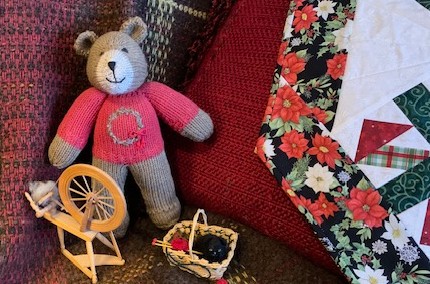 Knitted bear in a red sweater beside a tiny spinning wheel and a basket of yarn